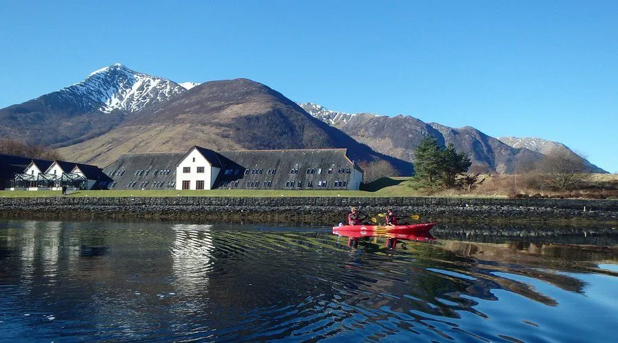 A scenic shot of two people in a red kayak on calm water, with a large, long white building and distant mountains visible under a clear blue sky.