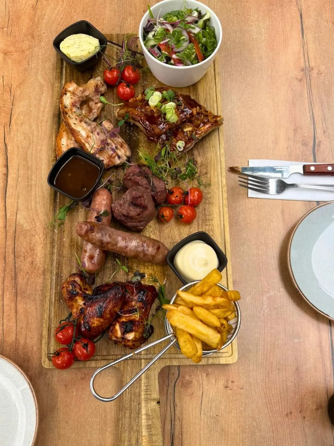 A well-lit, sharp, and photo-realistic image showcasing a large wooden platter filled with various grilled meats, including ribs, chicken, sausages, and steak, accompanied by french fries, cherry tomatoes, and small bowls of sauces.