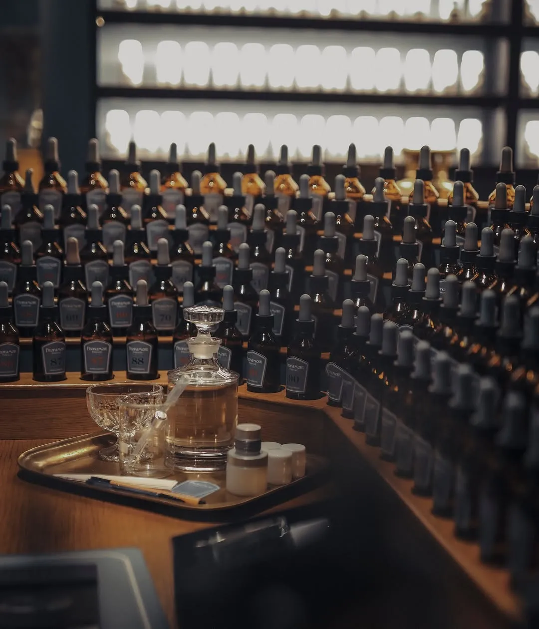 The image beautifully captures a collection of perfume or essential oil bottles, with a clear focus on a decanter and associated items arranged on a tray in the foreground. The depth of field effectively highlights the primary subject while creating an appealing backdrop of neatly organized bottles. The lighting is atmospheric, enhancing the visual appeal of the glass and liquid contents.