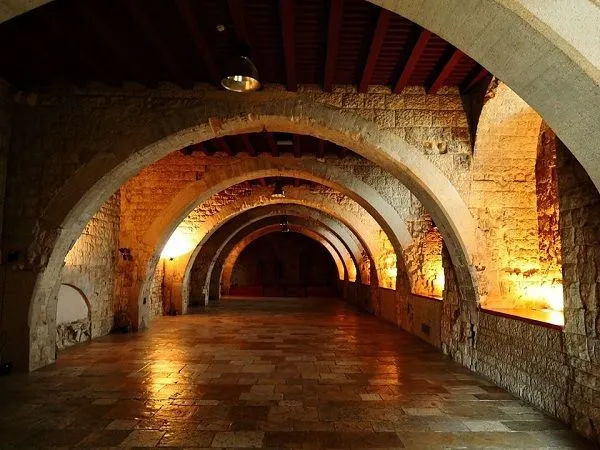 A long, vaulted stone hallway or corridor, illuminated by warm, directional lighting that highlights the arches and tiled floor.