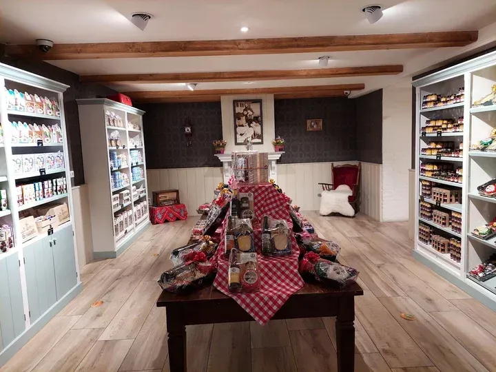 An interior shot of a gourmet food store or specialty shop, featuring a central display table with various packaged food products and shelves lining the walls. The room has a warm ambiance with wooden beams on the ceiling and light wooden flooring.