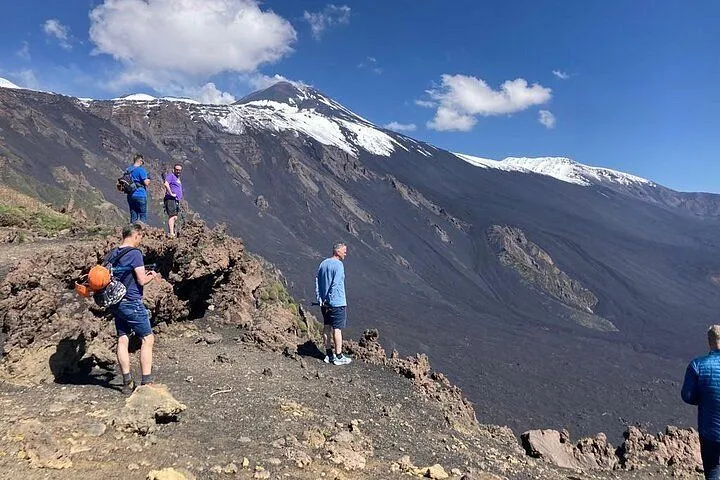 A group of people hiking on a rugged, dark volcanic landscape with a large snow-capped mountain in the background under a clear blue sky with some clouds.