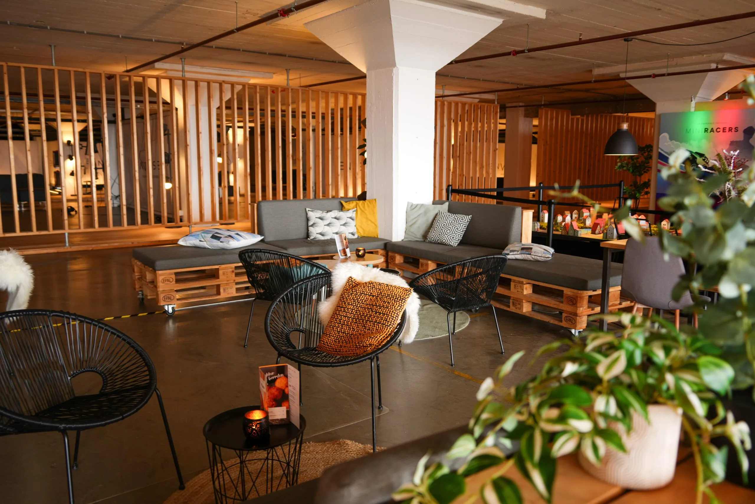 An image depicting a modern and cozy lounge area with wooden pallet seating, comfortable cushions, and stylish chairs. The space is well-composed with a warm, ambient lighting scheme.