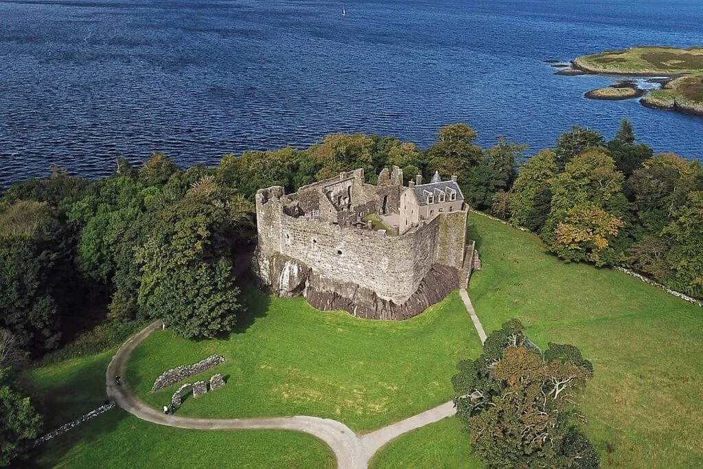 An aerial view of a historic castle situated by a large body of water, surrounded by green grass and trees. The image showcases the architectural details of the castle and its scenic environment.