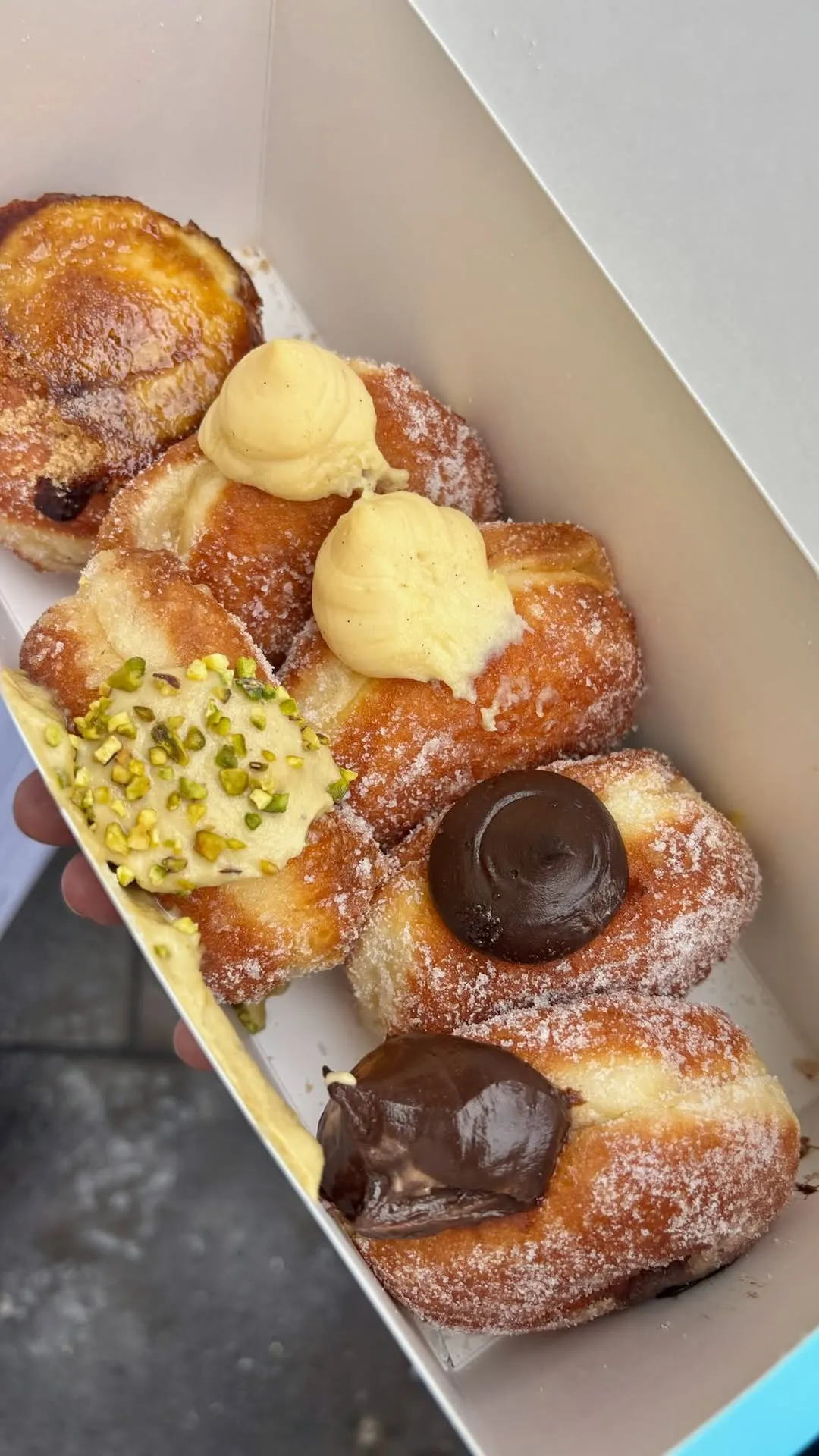 A close-up image of multiple sugar-dusted pastries, possibly choux buns or donuts, adorned with various toppings including vanilla cream, pistachio cream, and chocolate ganache, presented in a white box.