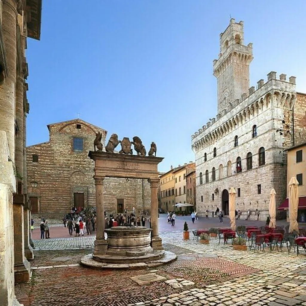 An image of a historic town square featuring a central well, traditional stone buildings, and a clear blue sky. The composition is balanced, capturing various elements of the square including people and outdoor seating areas.
