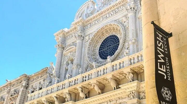 An editorial style image featuring a close-up view of an intricately carved Baroque church facade, likely made of light-colored stone, under a clear blue sky. The image highlights the detailed sculptures, columns, and a prominent rose window.