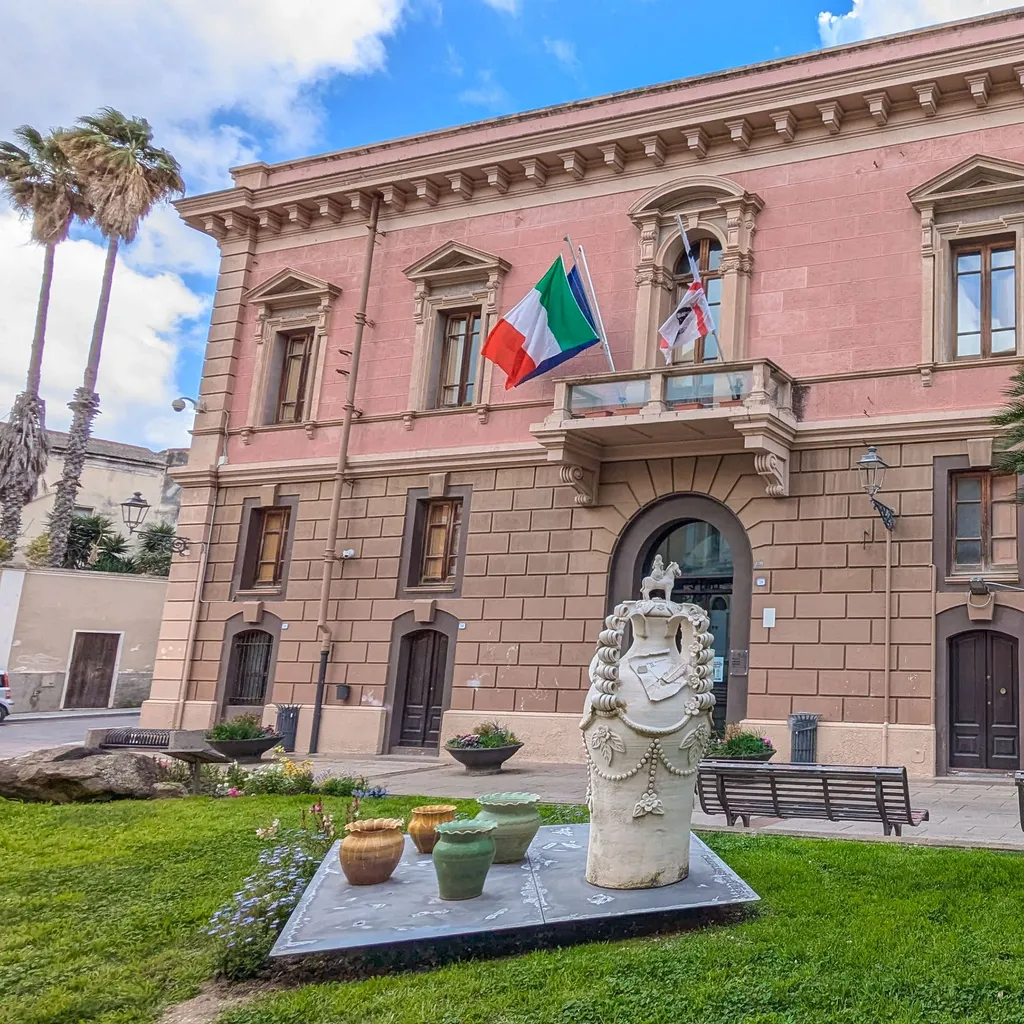 A well-composed and brightly lit photograph showcasing a pink historical building with an Italian flag, a prominent palm tree, and a decorative statue in the foreground. The image displays high clarity and natural colors.
