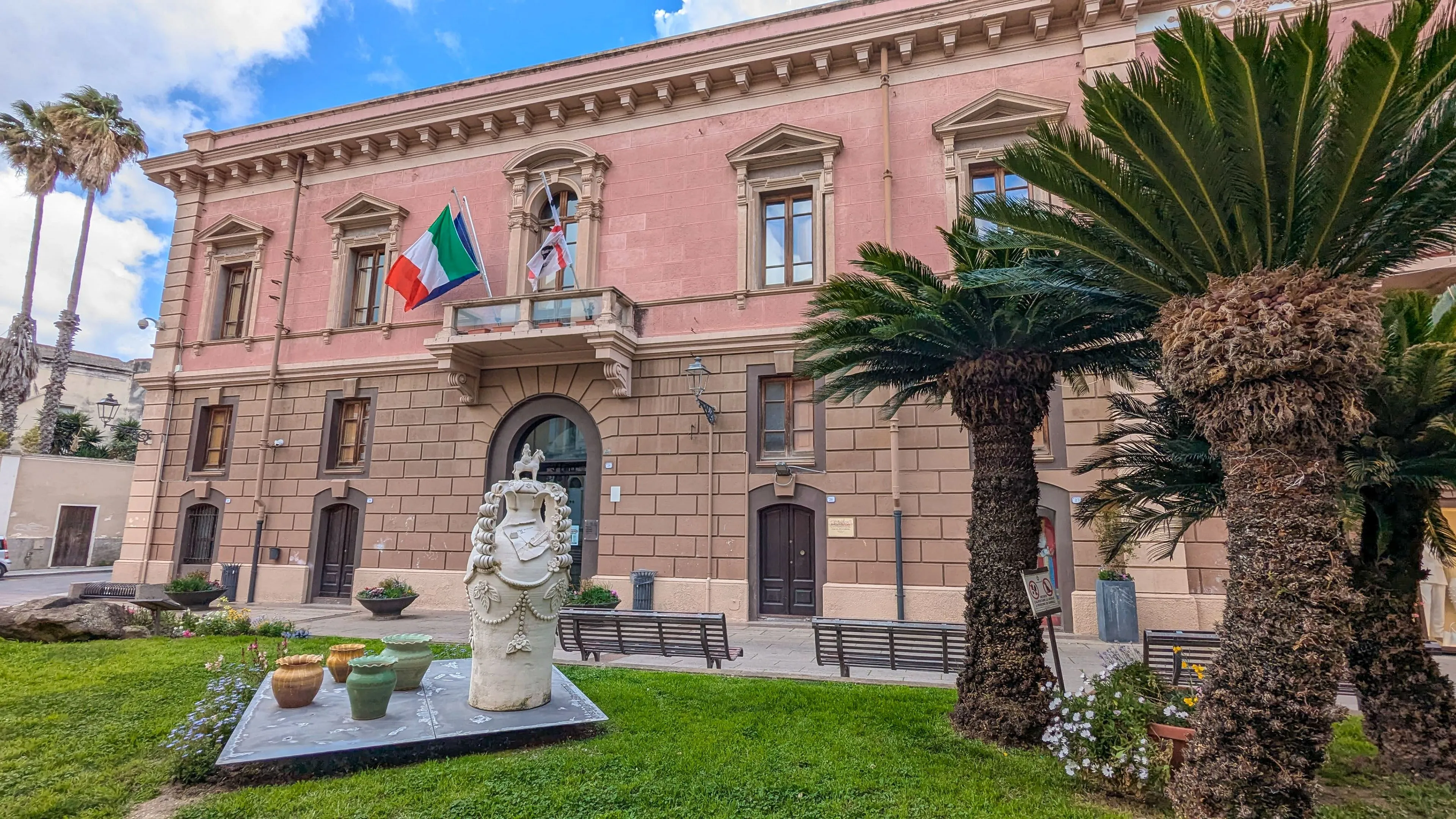 A well-composed and brightly lit photograph showcasing a pink historical building with an Italian flag, a prominent palm tree, and a decorative statue in the foreground. The image displays high clarity and natural colors.