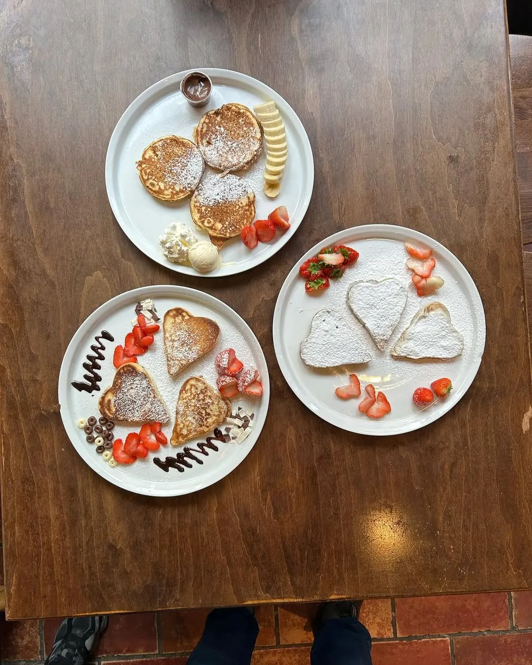 An overhead shot of three plates of pancakes, some heart-shaped, served with strawberries, bananas, powdered sugar, chocolate drizzle, and whipped cream on a wooden table.