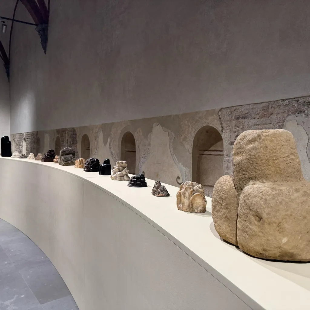 An interior shot of an exhibition space, featuring various sculptures or artifacts displayed on a curved white plinth. The background consists of an old stone wall with arched niches, characteristic of a historical building or museum.