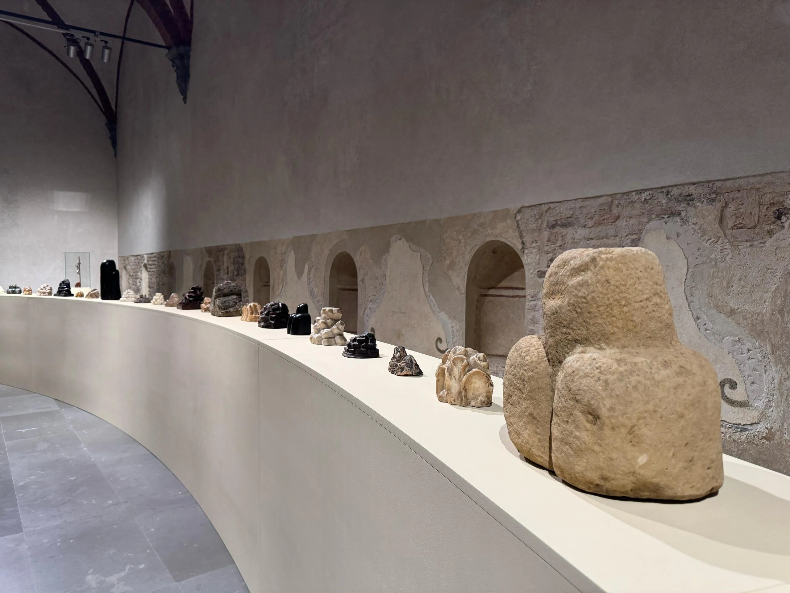 An interior shot of an exhibition space, featuring various sculptures or artifacts displayed on a curved white plinth. The background consists of an old stone wall with arched niches, characteristic of a historical building or museum.