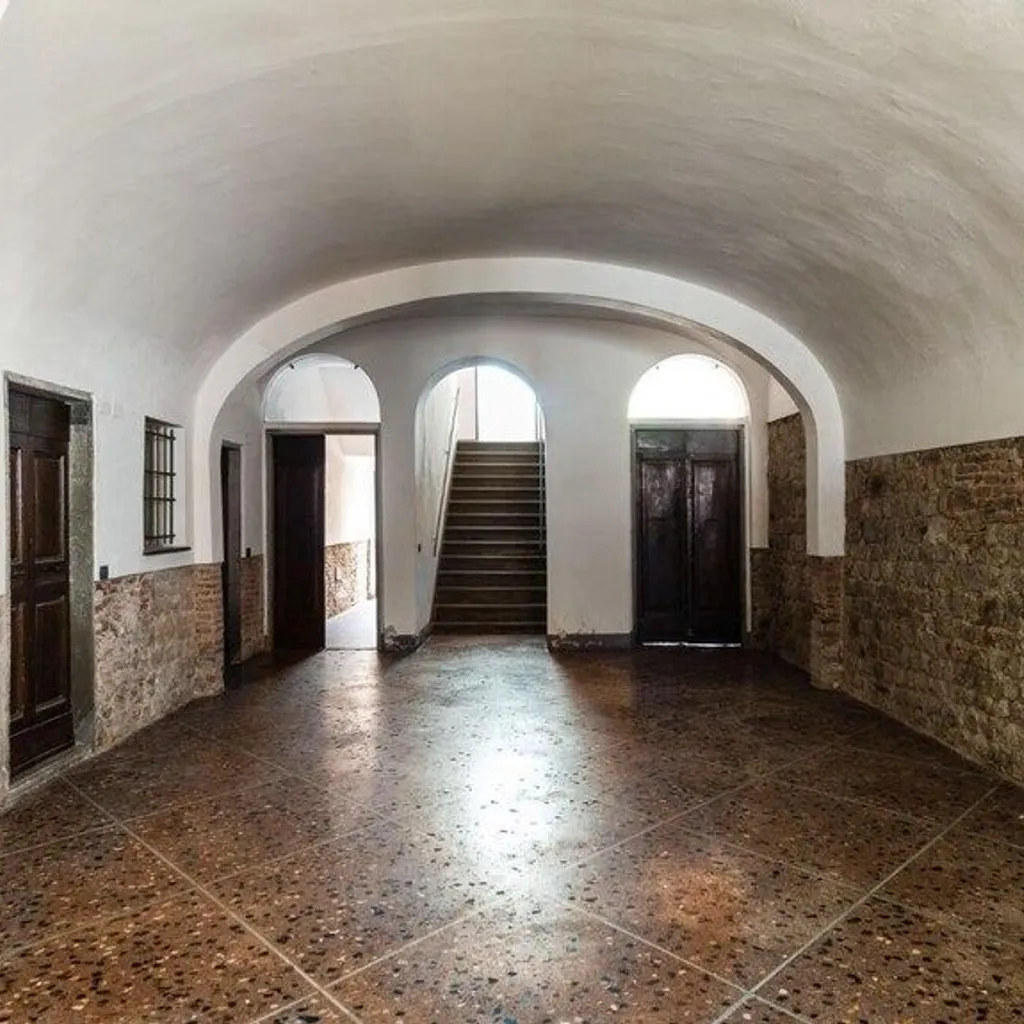An interior shot of an old building's hallway featuring a vaulted ceiling, brick and stone walls, and a visible staircase at the end. The foreground has a patterned floor.