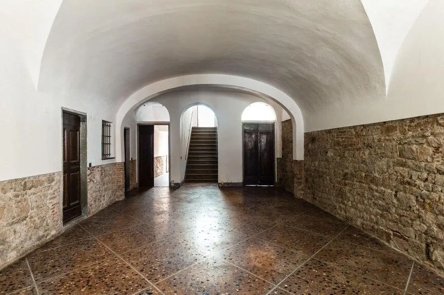 An interior shot of an old building's hallway featuring a vaulted ceiling, brick and stone walls, and a visible staircase at the end. The foreground has a patterned floor.