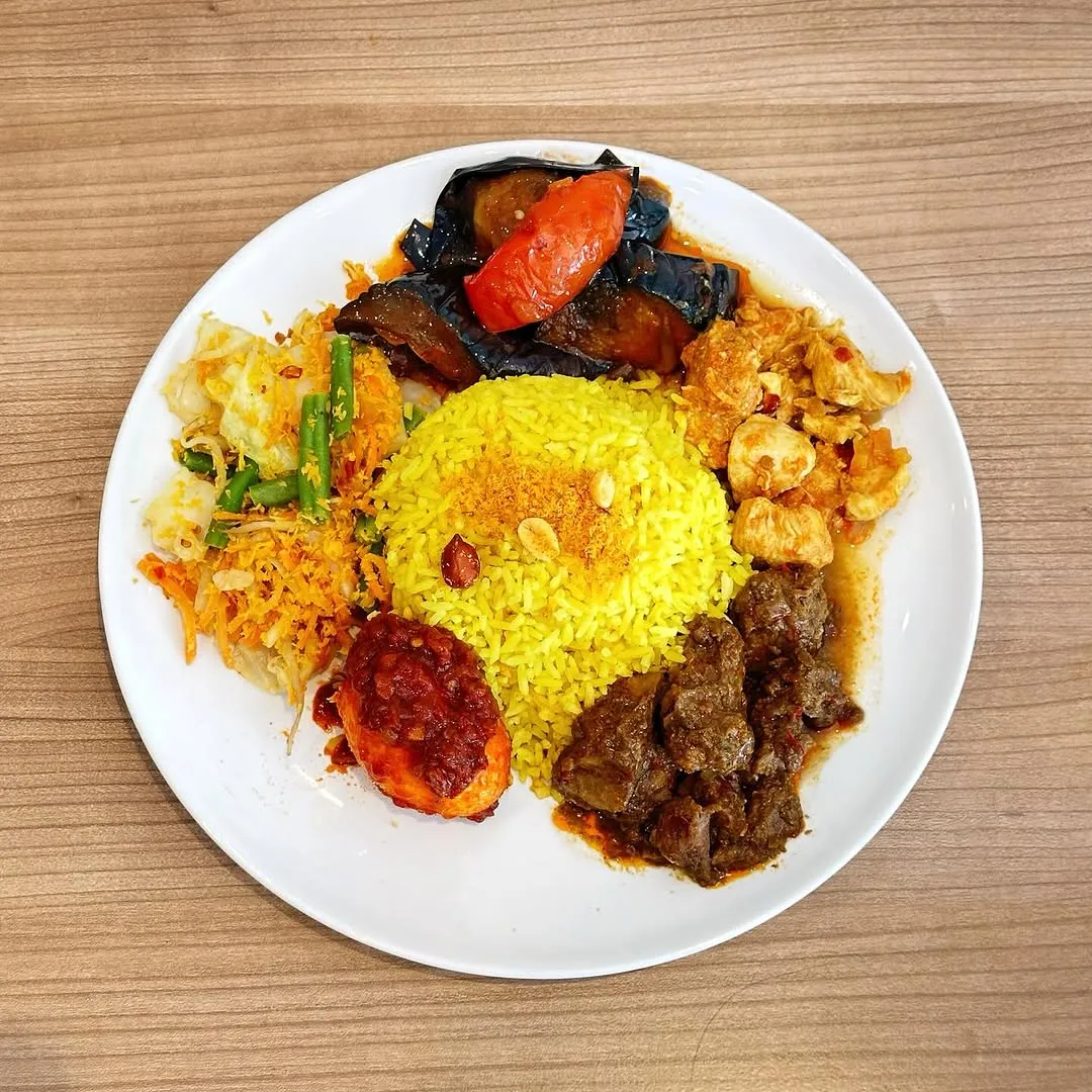 A vibrant close-up shot of an assorted meal on a white plate, featuring yellow rice surrounded by various side dishes including eggplant, meat, egg, and mixed vegetables. The meal is presented on a clean wooden table.