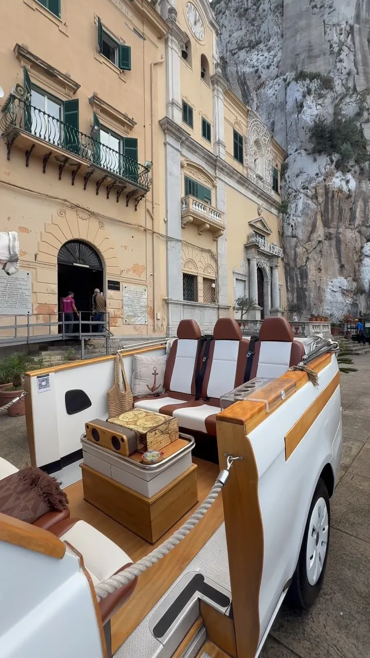 A unique custom-built open-top vehicle featuring brown and white seats, wooden trim, and a small table with decorative items, parked on an outdoor street or plaza. In the background, there is an old light-yellow building with balconies and a natural rock formation.