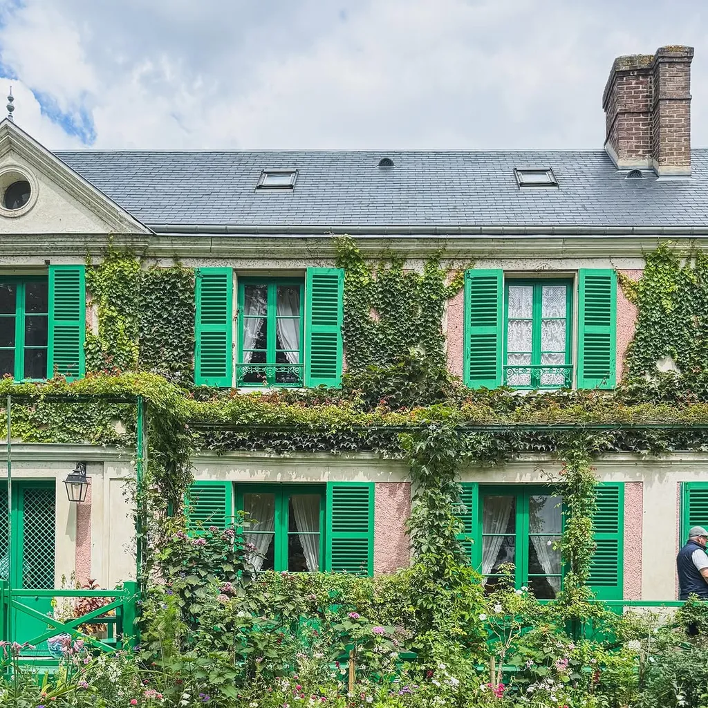 A vibrant image showcasing a charming house with green shutters and abundant ivy, surrounded by a lush, colorful garden with various flowers and a winding gravel path. A green bench is visible on the right.
