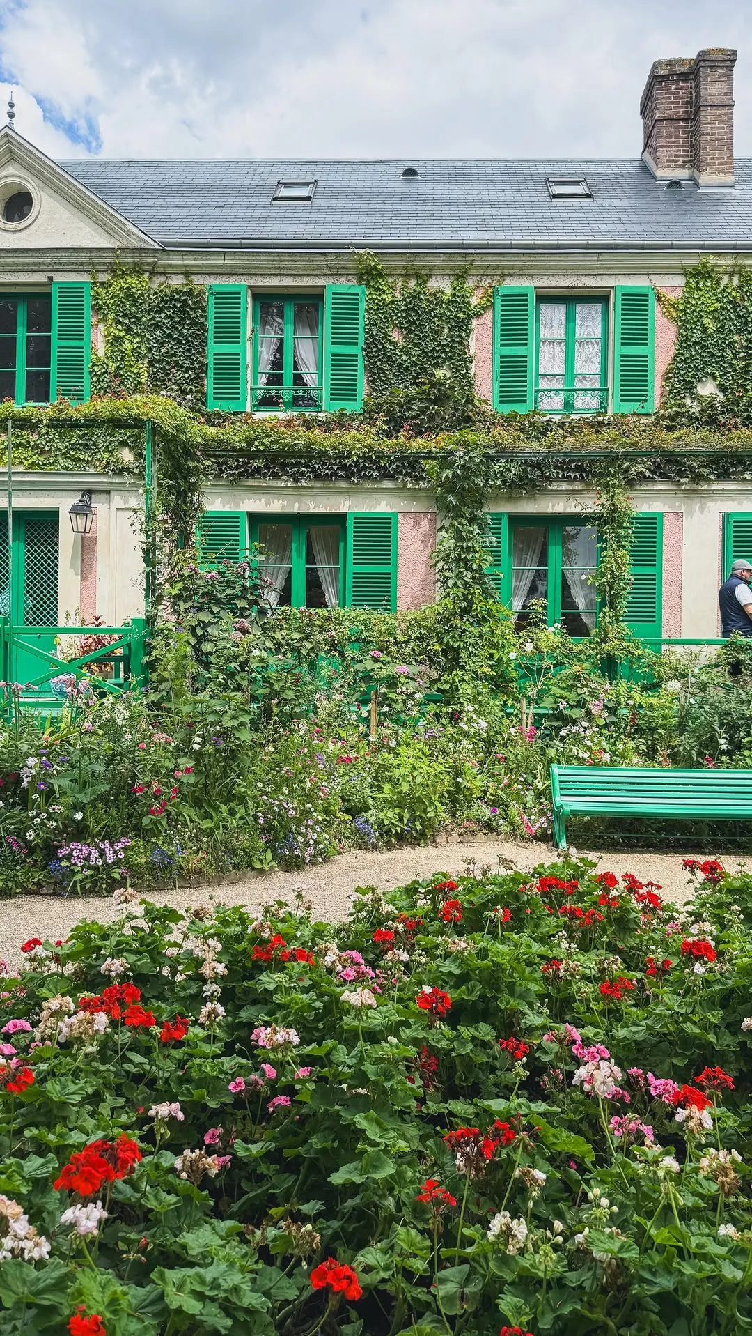 A vibrant image showcasing a charming house with green shutters and abundant ivy, surrounded by a lush, colorful garden with various flowers and a winding gravel path. A green bench is visible on the right.