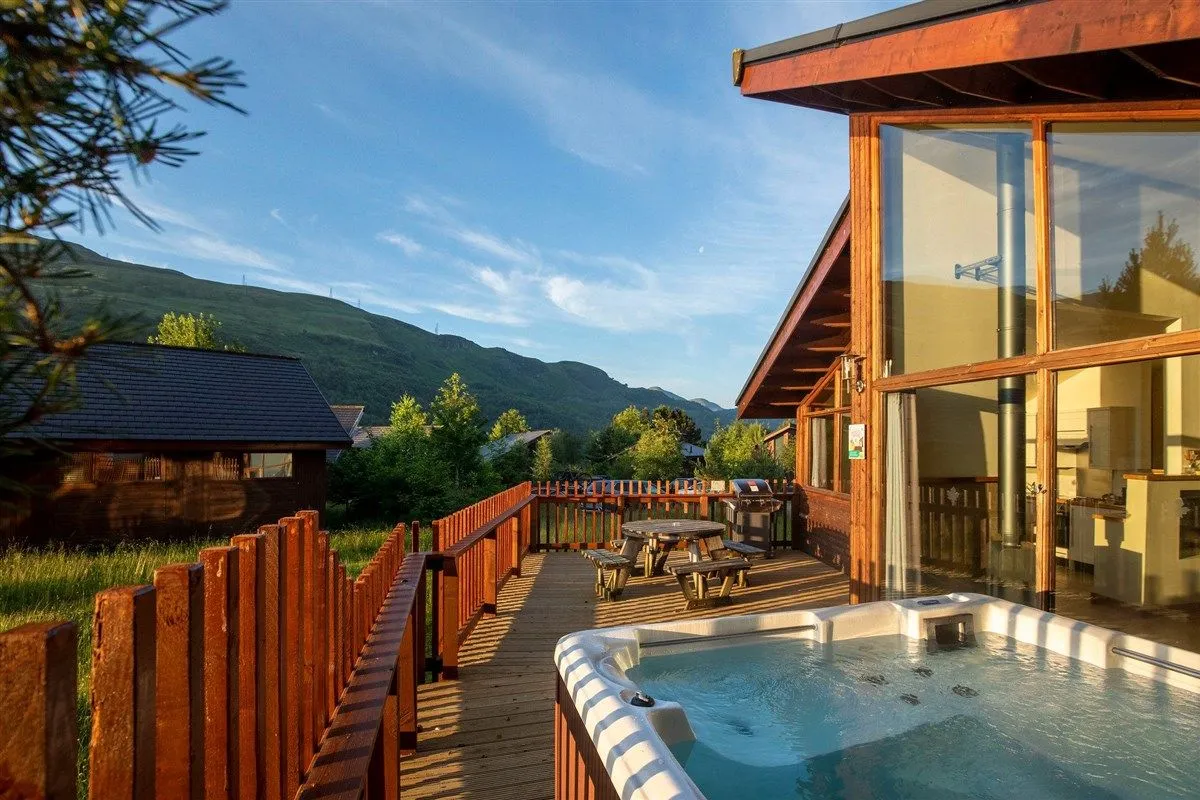 The image captures a wooden lodge deck featuring a hot tub and a picnic table, set against a backdrop of mountains under a clear sky. The composition highlights the outdoor amenities of the accommodation with a pleasant scenic view.