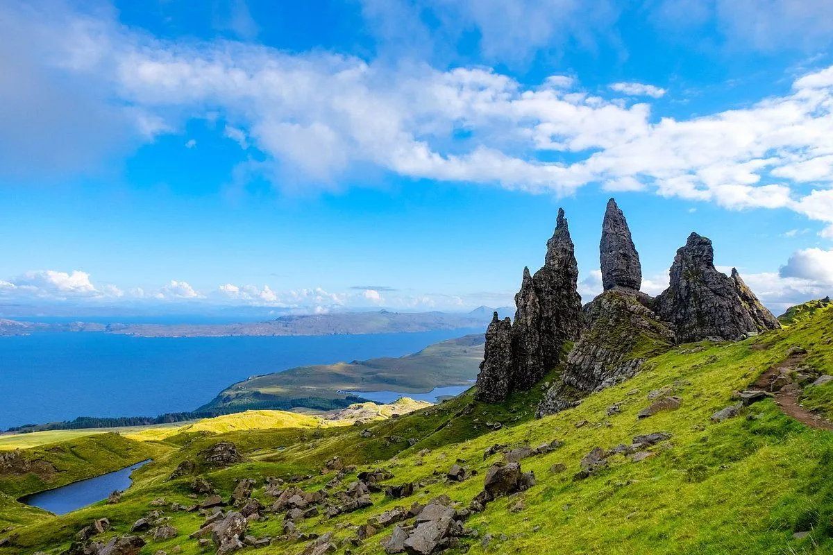 A vibrant and well-composed landscape photograph featuring the iconic Old Man of Storr rock formations on the Isle of Skye, Scotland, with lush green hills, a large body of water, and a bright blue sky with white clouds.