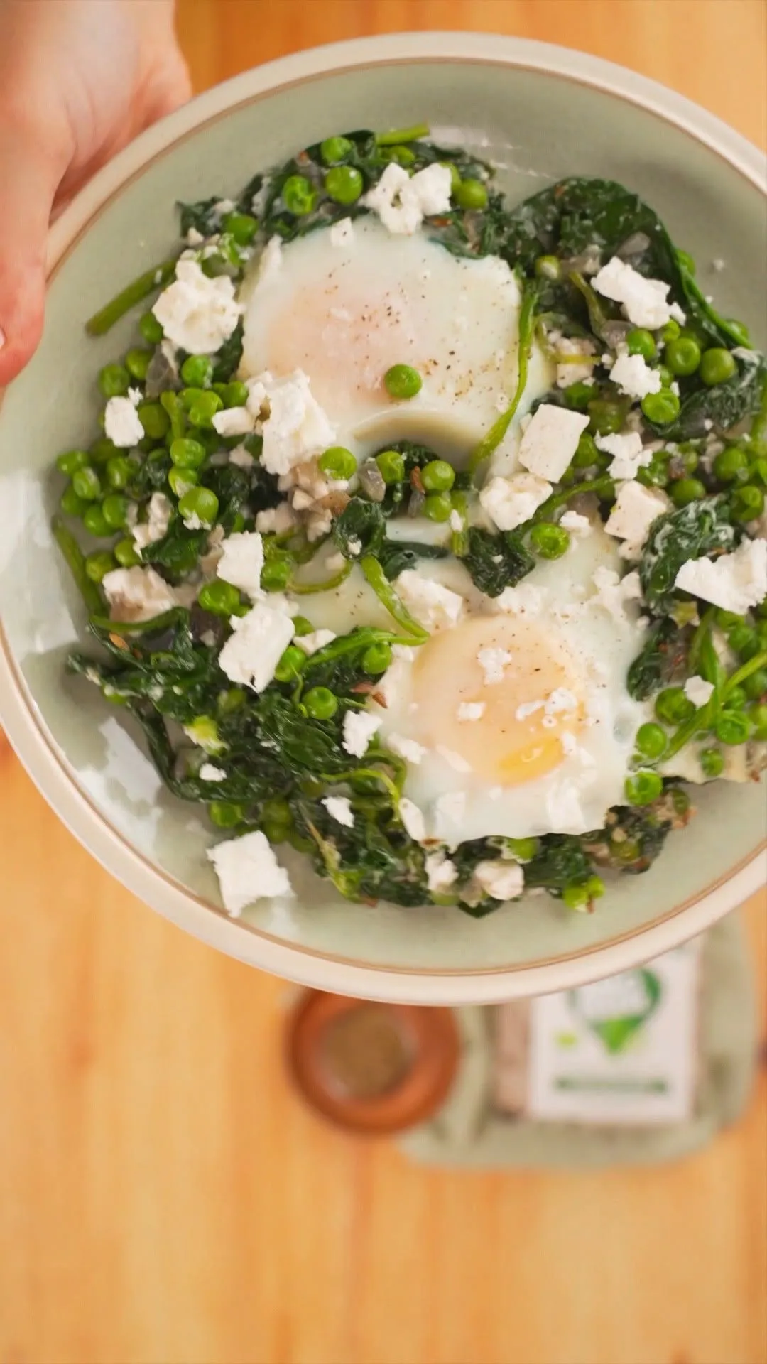 A top-down view of a dish featuring two fried eggs nestled among vibrant green spinach and peas, topped with crumbled white feta cheese.