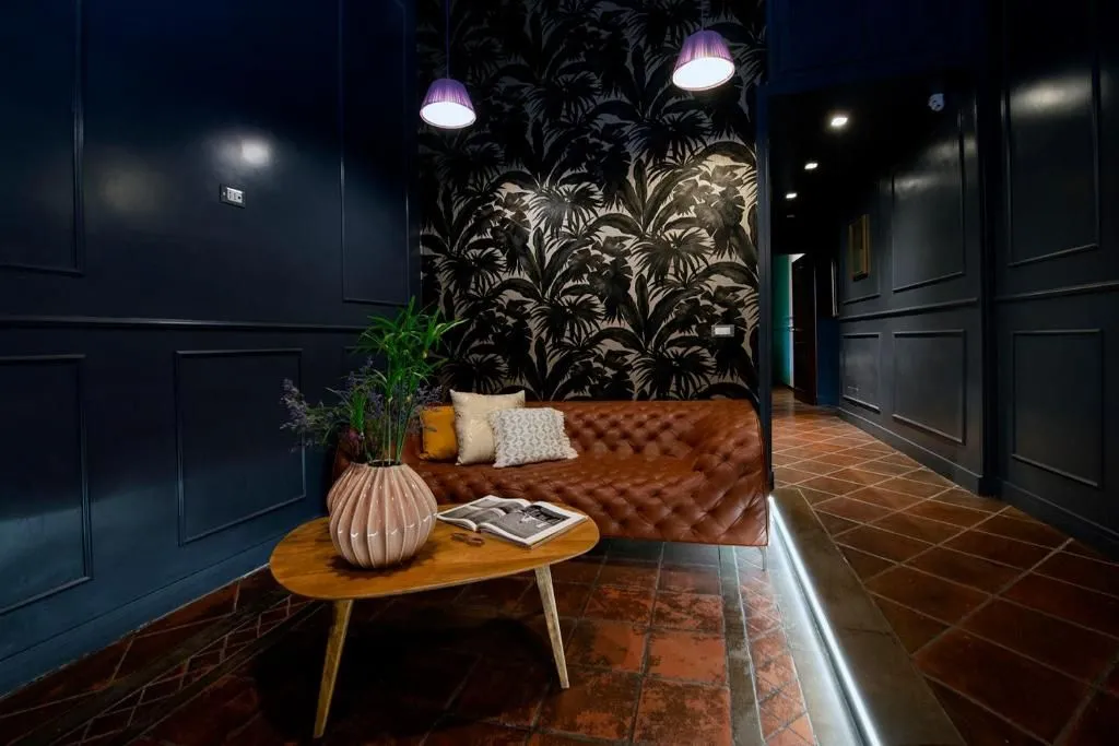 An interior shot featuring a brown leather tufted sofa with throw pillows, a wooden coffee table with a vase and a book, against a dark wall with tropical-patterned wallpaper. The room extends into a dimly lit hallway with tiled flooring and a strip of floor lighting.