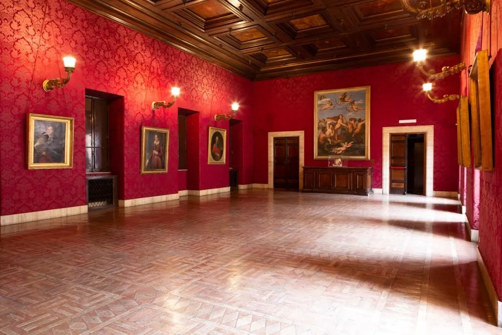 The image effectively captures a grand, ornate interior room, highlighting the vibrant red textured walls, patterned tiled floor, and detailed wooden coffered ceiling. It is sharp and photo-realistic, with most key elements in focus, allowing for clear appreciation of the architectural and decorative details. However, the lighting is uneven, with noticeable glare from the wall sconces creating bright spots and some areas appearing slightly underexposed, impacting overall clarity and balance. The composition is decent but could be more dynamic.
