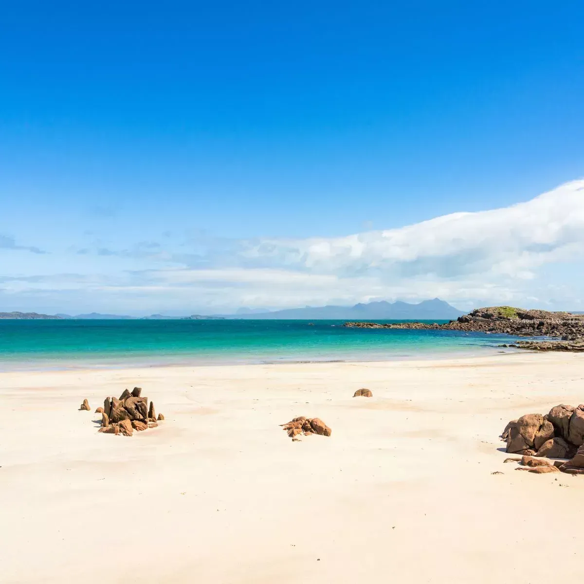 This is an exceptionally well-composed and vibrantly lit image of a pristine beach. The colors are striking, particularly the clear blue sky and turquoise water, appearing highly color-accurate and photo-realistic. The image is tack sharp from the foreground rocks and sand to the distant mountains, with all key elements perfectly in focus. The overall quality is editorial.