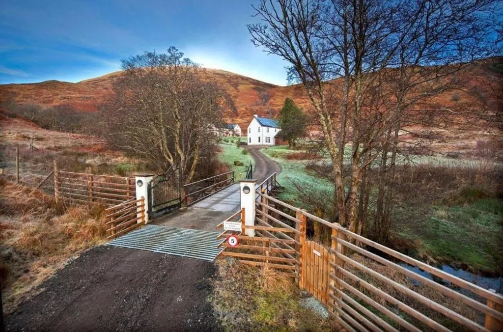 A picturesque landscape featuring a white house nestled in a valley, accessed by a dirt road and a bridge with a cattle grid, surrounded by hills and bare trees under a clear sky.