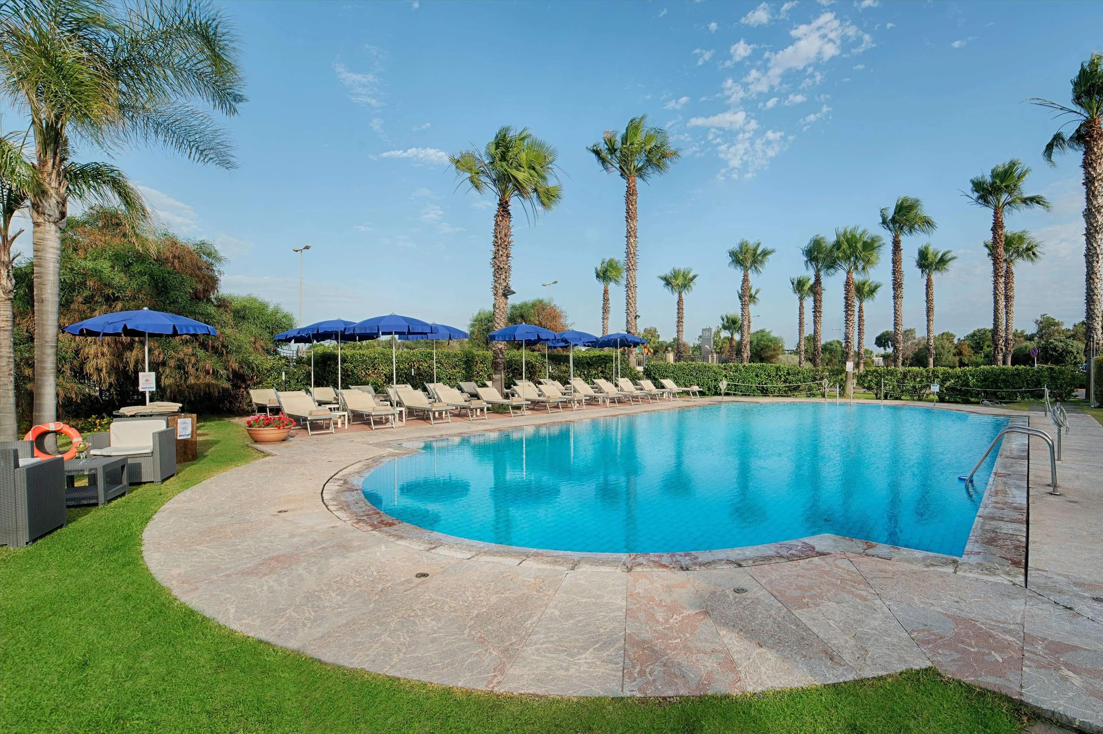 An inviting outdoor swimming pool surrounded by lounge chairs and palm trees under a clear blue sky. The scene evokes a sense of relaxation and tropical vacation.