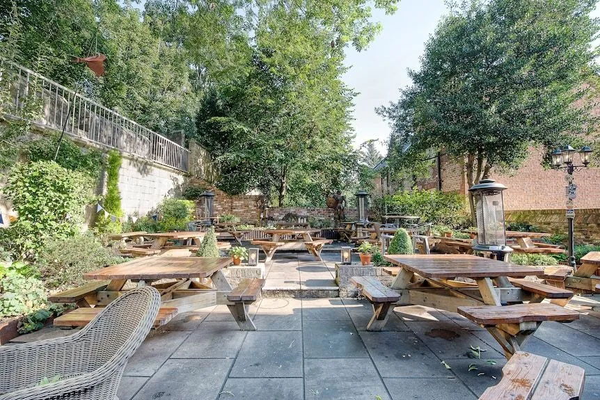 An outdoor patio area with multiple wooden picnic tables and benches, surrounded by lush green trees and shrubs, under natural daylight. The paving is grey stone, and there are brick walls and a metal railing in the background.