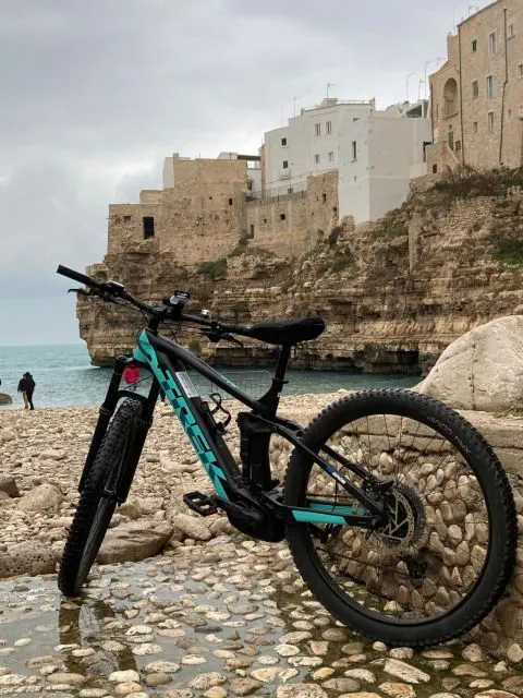 An e-bike rests on a rocky beach with historical white buildings perched on cliffs overlooking the sea in the background, under an overcast sky.