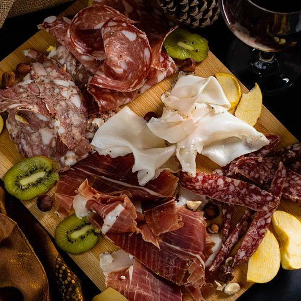 An overhead shot of a charcuterie board featuring a variety of sliced cured meats, including salami and prosciutto, alongside sliced kiwi, apple wedges, and scattered nuts. A dark background with a visible wine glass in the upper right corner sets a cozy and inviting atmosphere.