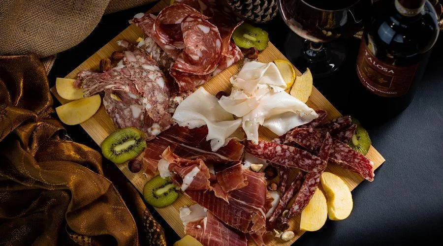 An overhead shot of a charcuterie board featuring a variety of sliced cured meats, including salami and prosciutto, alongside sliced kiwi, apple wedges, and scattered nuts. A dark background with a visible wine glass in the upper right corner sets a cozy and inviting atmosphere.
