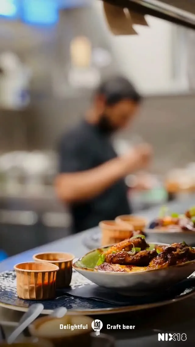 A close-up photograph of several dishes, possibly chicken wings or similar appetizers, garnished and presented on plates, with small serving cups alongside. The background is blurred, showing a person working in a kitchen or food preparation area.