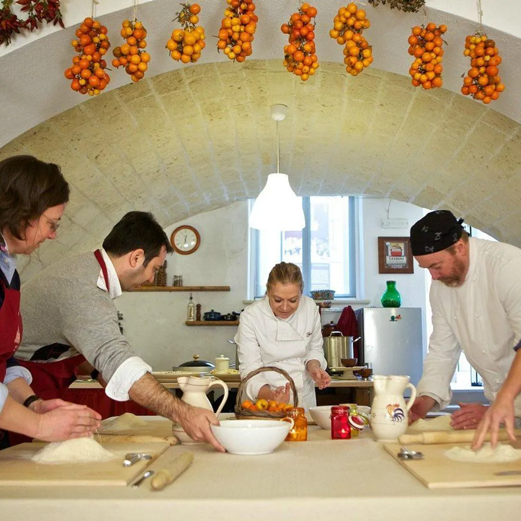 An editorial-style image depicting a cooking class or workshop. Several individuals, including a chef, are gathered around a table, actively engaged in preparing food, likely pasta, using ingredients like flour. The scene is set in a rustic kitchen with an arched ceiling adorned with hanging bundles of small, ripe tomatoes, adding to the authentic atmosphere.