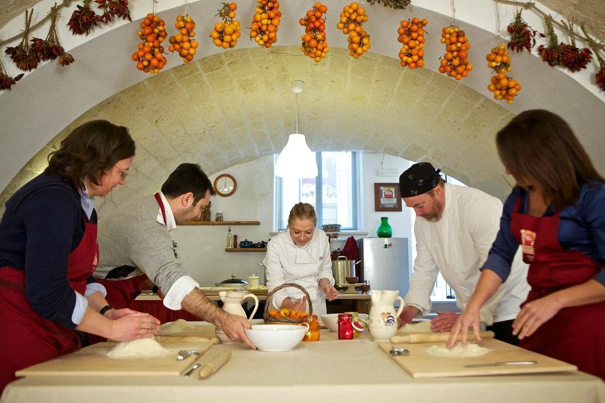 An editorial-style image depicting a cooking class or workshop. Several individuals, including a chef, are gathered around a table, actively engaged in preparing food, likely pasta, using ingredients like flour. The scene is set in a rustic kitchen with an arched ceiling adorned with hanging bundles of small, ripe tomatoes, adding to the authentic atmosphere.