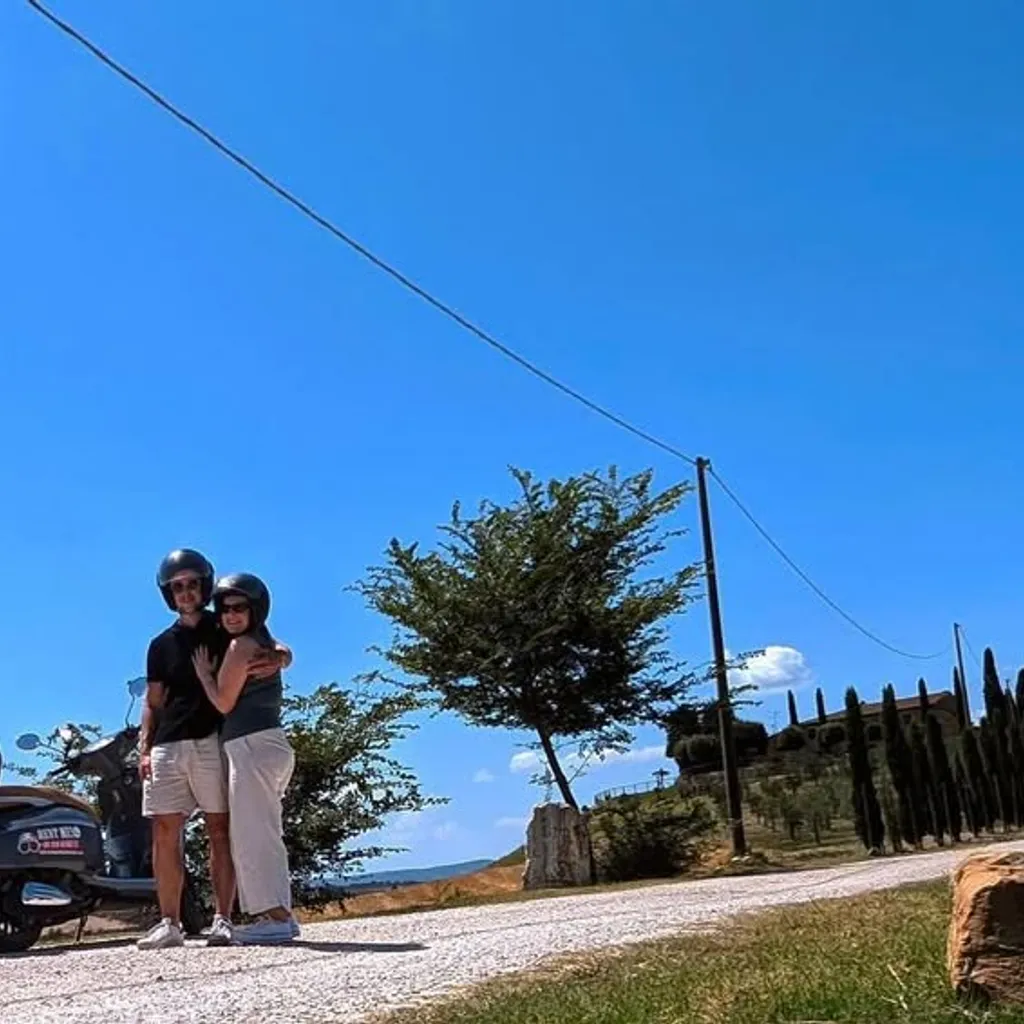A bright, sunny image of a rural landscape, likely in the countryside, featuring a dirt road winding through fields, distinct trees, and a clear blue sky. A power line and pole are visible in the upper portion of the image.