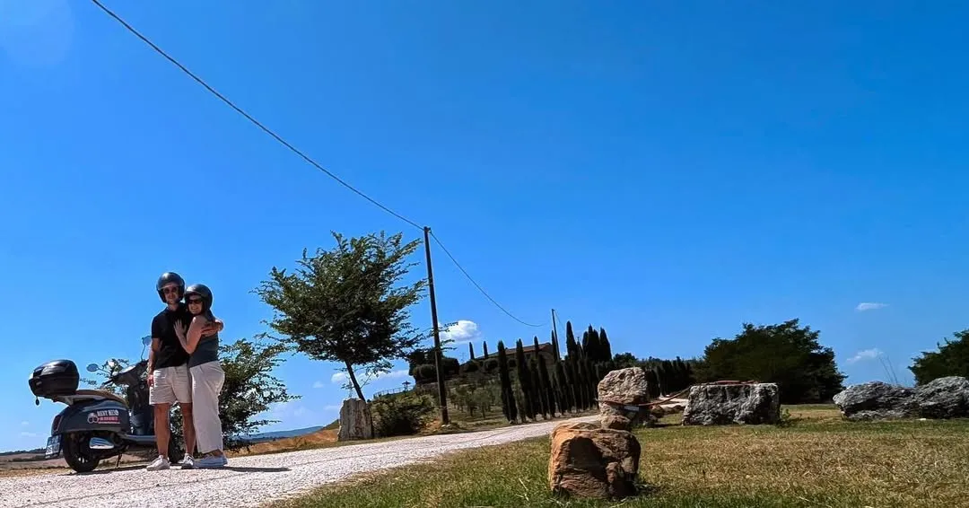 A bright, sunny image of a rural landscape, likely in the countryside, featuring a dirt road winding through fields, distinct trees, and a clear blue sky. A power line and pole are visible in the upper portion of the image.