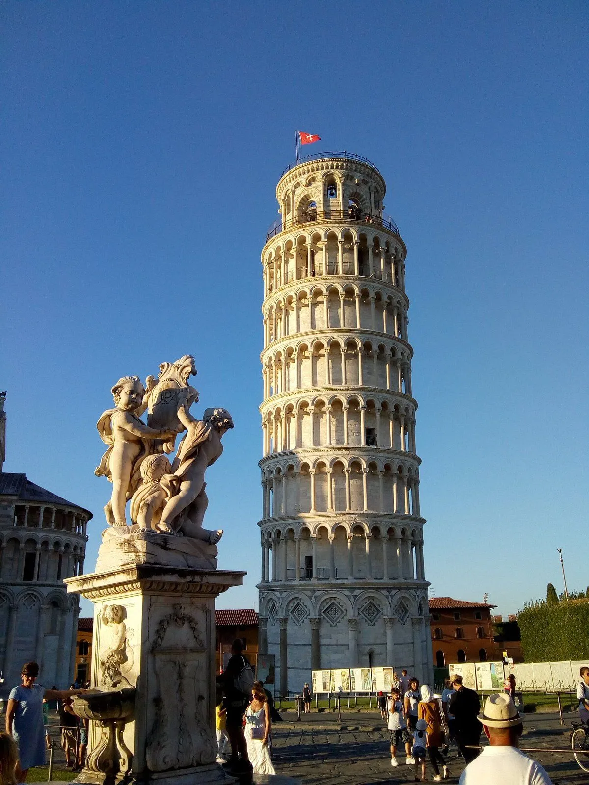 The image prominently features the Leaning Tower of Pisa and a classical statue against a clear blue sky. The composition effectively highlights the iconic landmark and the foreground sculpture, both rendered with excellent sharpness and detail. The natural lighting is superb, illuminating the stone structures and providing accurate, vibrant colors. The image feels authentic and photo-realistic, with all key elements in clear focus.