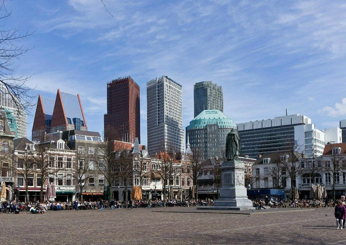A well-composed and photo-realistic image depicting a bustling city square with a blend of historic and modern architecture. The scene is well-lit under clear daylight, showcasing accurate colors and generally sharp details across the key elements. The image successfully captures the dynamic atmosphere of an urban public space.