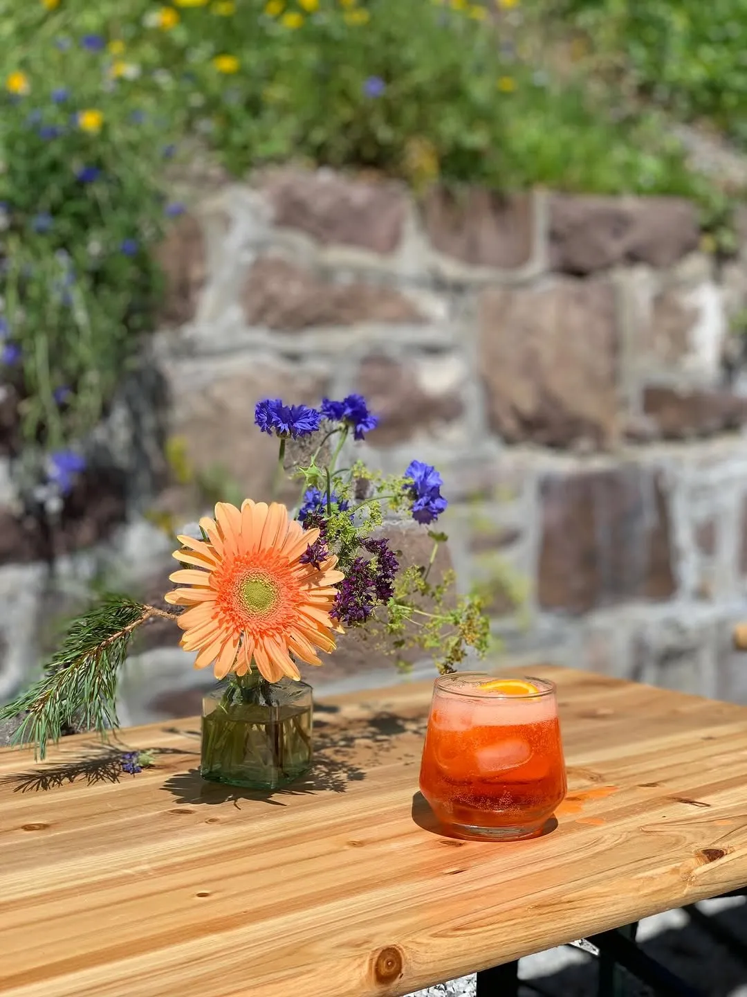 The image features a well-composed shot of a vibrant orange drink alongside a flower arrangement on a wooden table, with a stone wall background. The natural lighting is good, creating bright subjects and pleasant colors. Key elements like the drink and the flower are sharp and in focus, contributing to a photo-realistic and appealing visual.