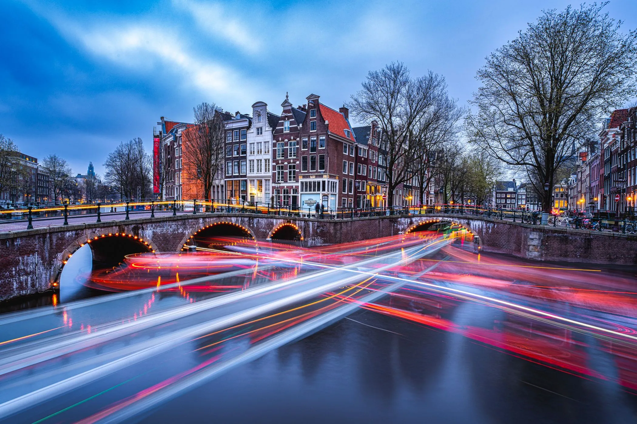 An exquisite long exposure photograph of a canal in Amsterdam at blue hour, featuring historic gabled buildings, arched bridges, and dynamic light trails from passing boats on the water.