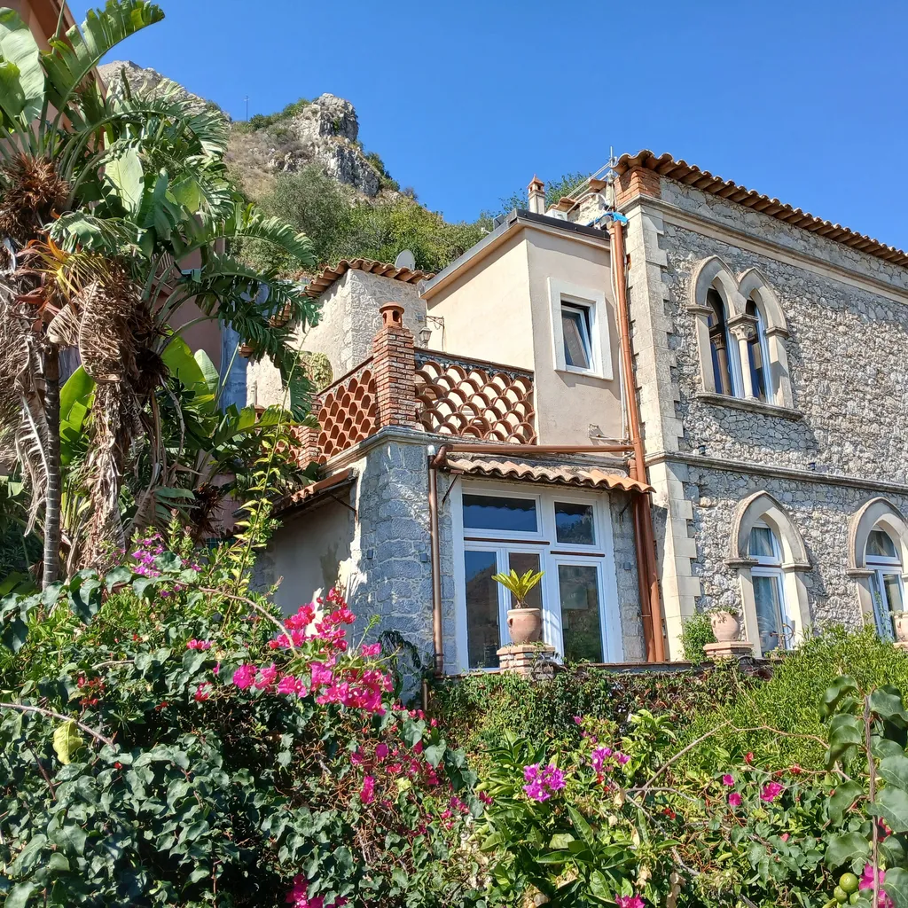 An editorial style image showcasing a charming stone villa nestled amidst lush green foliage and vibrant pink flowers, with a mountain backdrop. The image benefits from excellent natural lighting, sharp details, accurate colors, and a strong sense of photo-realism. The composition is generally well-executed, with key elements in focus.