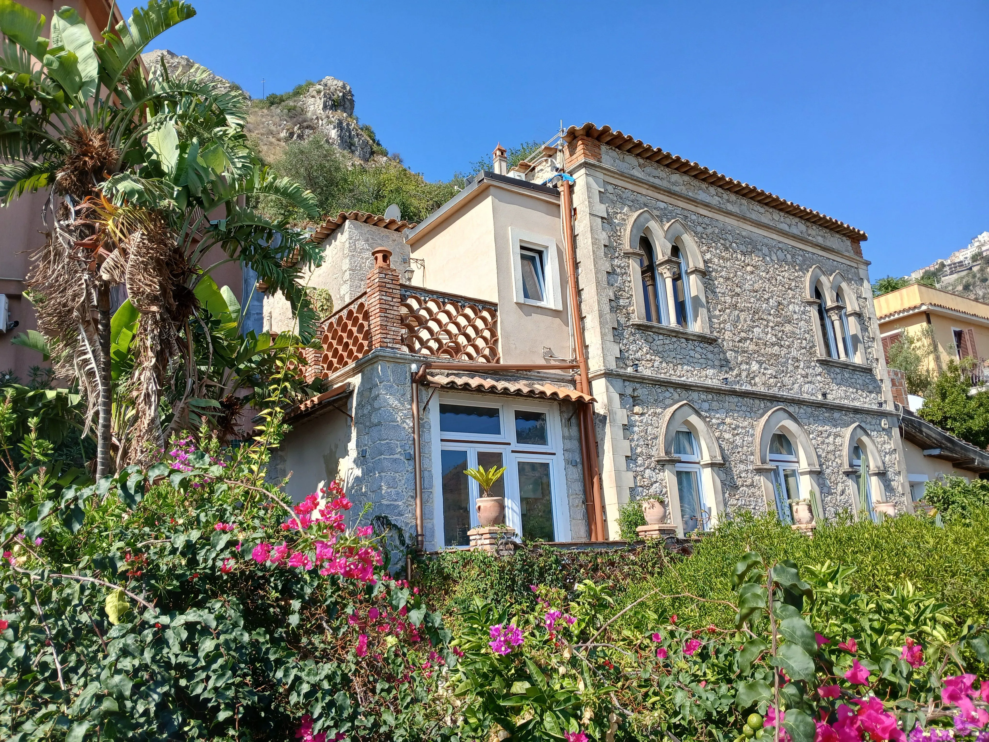 An editorial style image showcasing a charming stone villa nestled amidst lush green foliage and vibrant pink flowers, with a mountain backdrop. The image benefits from excellent natural lighting, sharp details, accurate colors, and a strong sense of photo-realism. The composition is generally well-executed, with key elements in focus.