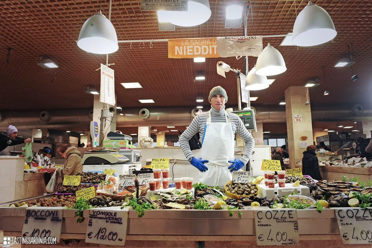An editorial image of a male vendor at a bustling seafood market stall. The vendor, wearing an apron and gloves, stands behind a display of fresh seafood, including mussels and clams, with various price signs visible. The market environment is well-lit by overhead lights, and other shoppers are faintly visible in the background.