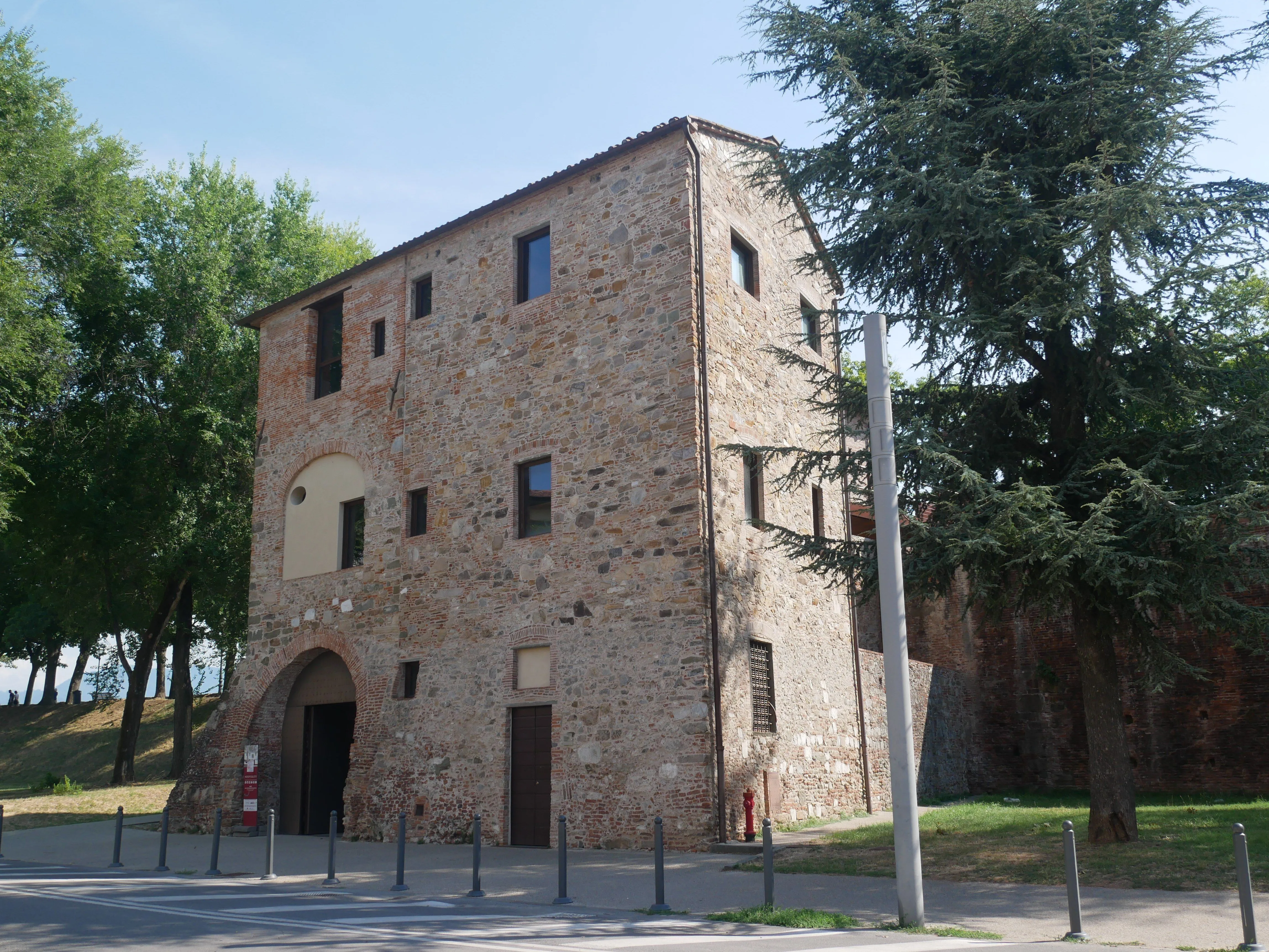 A photograph of an old stone and brick building under a clear sky, with trees on either side and street elements in the foreground.