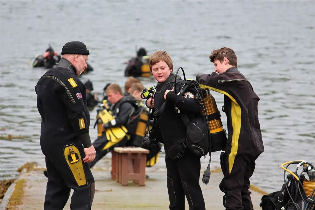 The image is well-composed, focusing on the interaction between two divers preparing their gear. The main subjects are sharp and in focus, contributing to the photo-realistic quality. Colors appear natural and accurate for the outdoor setting. The lighting, while sufficient, is somewhat flat, lacking strong highlights or shadows, which slightly diminishes the overall visual impact.