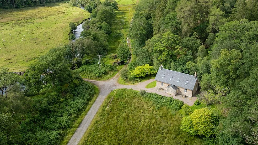 An aerial view of a stone cottage nestled in a lush green valley, surrounded by dense trees, a winding river, and dirt roads.