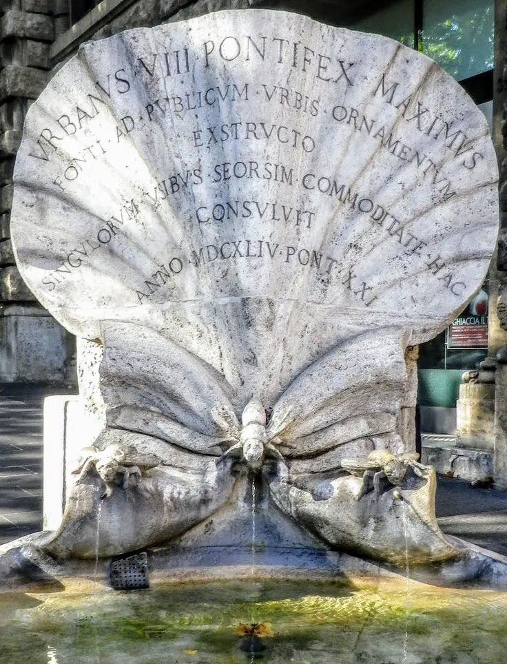 An editorial-style close-up photograph of a historic stone fountain, possibly in Rome, featuring a large shell-shaped backplate with Latin inscriptions and water spouting from intricately carved insect-like figures into a lower basin. The natural light illuminates the stone, highlighting its texture and details.